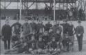 a black and white photograph of the Football team of 1893 in front of the grandstand on the athletic field. the football team is standing in rows on the field and spectators stand in the grandstand behind them.