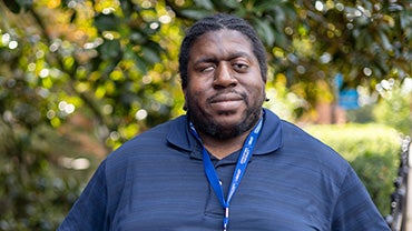 A portrait of Kali Bundu, a middle-aged Black man standing in front of trees.