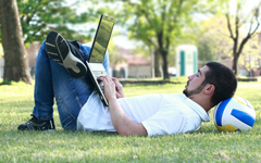 Male student working outside at a laptop while lying on the ground with his head on a volleyball.
