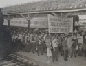 a black and white photograph of a marching band standing on the platform of a train station beneath a banner that reads, "georgetown alumni of greater miami welcomes georgetown men"