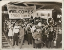 A black and white photograph of the Georgetown University Band in Miami for the Orange Bowl game. The band stands on the platform of the train station under a banner that reads "Georgetown Alumni of Greater Miami Welcomes Georgetown Men"