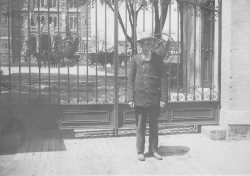 Black and white photograph of a bearded man standing in front of the closed Georgetown College gates