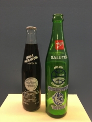Color photograph of a full commemorative Coke bottle and an empty commemorative 7 UP bottle