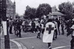 Black and white photograph student medic wearing a white cape with a cross painted on it walking towards a crowd at the main gates