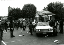 Black and white photograph of protesters outside the main gates, with a white sedan parked in the intersection