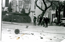 Black and white photograph of police in riot gear standing next to debris in the street