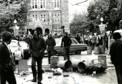 Black and white photograph of protesters on O Street east of the main gates, with a line of trash cans blocking the street