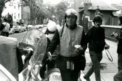 Black and white photograph of a policeman eating a sandwich while leaning on his scooter, near the Prospect entrance of the Car Barn, with several photographers in the background