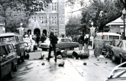 Black and white photograph of protester crossing O Street, with a line of trashcans behind him and the crowd of people at the main gates