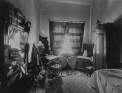 Black and white photograph of a dorm room including a bed, dresser and wardrobe