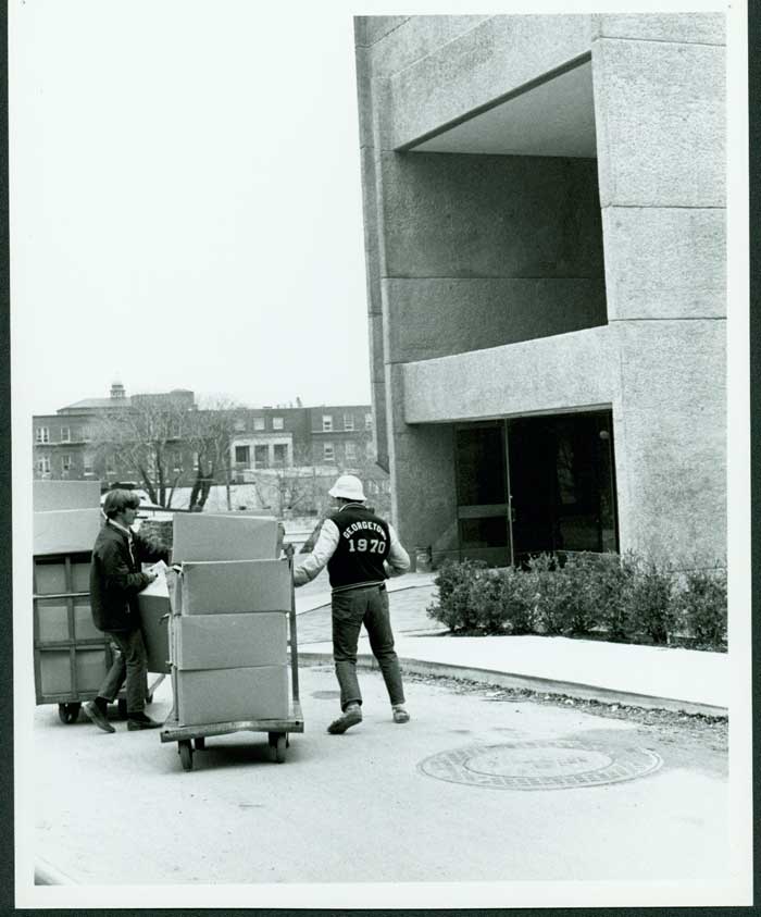 Univeristy staff push carts full of books in boxes towards the entrance of Lauinger Library