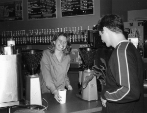 Image of a Corp member behind the counter at Uncommon Grounds. She is smiling and handing a male student a cup.
