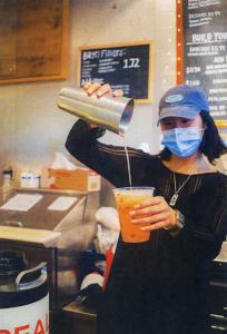 Corp member Nina pours a drink for a customer while at work in the Midnight Mug. She is wearing a hat and a face mask and pours liquid from a silver shaker in her right hand into a plastic cup in her left hand.