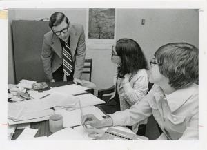 In this black and white photograph, three people sit around a table covered in papers.
