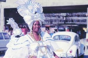 Color photo of a middle aged woman wearing a costume that is big white dress, gold beads, and a white headpiece. Photo includes her side profile, body from the torso up, and a white car in the background.