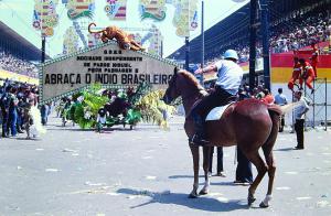 Color photo of officer on horse in front of a large, tropical float. The officer has extended his arm, only the back of his body and a semi profile of the horse are visible.