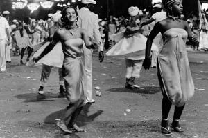 Blurry black and white photo of people wearing white and dancing in the street. The two main subjects are young women. One smiles and is turned towards the camera and the other (to the right) is looking away, focused on her dance.