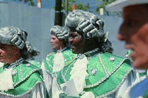Color photo of men dressed up as French nobility in a carnival event. They are in profile, and the costumes include white, green, and a gray wig.