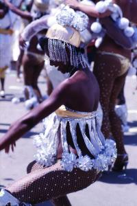 Color profile photo of a woman crouched in a dance during carnival. She wears a costume of a headpiece, sparkly frayed top, and tights.