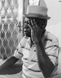 Black and white portrait of an older man sitting in semi profile. He wears a striped polo shirt and holds one hand to his temple.
