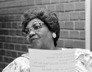 Black and white headshot of a woman (presumably standing) in front of a brick wall and wearing glasses. She holds a piece of paper in front of her chest, but tilts her head to the side.