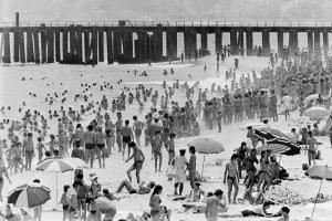 Black and white photo of a stretch of Ipanema Beach full of swimmers and people on the sand. There are some beach umbrellas and a pier in the background.
