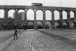 Black and white photo of a man walking on cobblestones in front of a towering old aqueduct