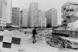 Black and white photo of a man standing in a construction site. He is turned away from the camera towards a decrepit building and some of Rio's skyline.