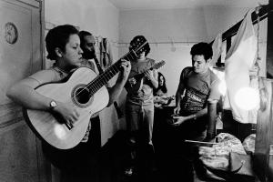 Black and white photograph of a group of four men playing different instruments inside a small room. The most forward man is in profile and playing the guitar.