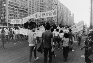 Black and white photo of the backs of people holding banners and walking in a street parade.