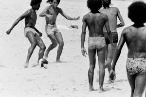 Black and white image of five full bodied shirtless men playing a game of soccer.