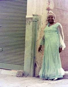 Color photographic portrait of woman standing in front of building corner. She wears a green long sleeve dress which echoes the green tint of the closed shop or garage around the corner.