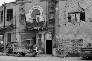 An old man with a cane sits in front of buildings that are cracked and damaged