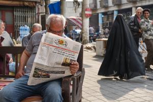 An older man reads a newspaper. In the background we see the back of a woman in traditional Iraqi clothing and two men look directly at the camera. The men are smiling. 