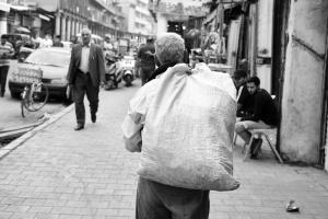 Black and white photograph of an older man walking away from the camera. He is carrying a large fabric sack slung over his back. In the background is a busy street with people, cars, a bike, and a motorcycle.