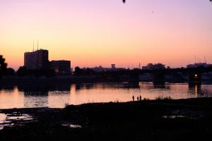 Color photo of a sunset with gradient of color from purple to yellow to pink in the sky that is reflected in the water of the river. Children can be seen along the edge of the river in the distance. A city with buildings of varying heights is on the opposite side of the river.