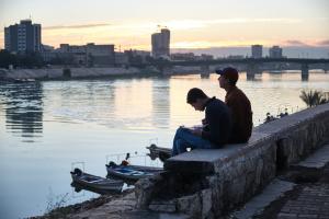 A color photograph of two boys sitting along the banks of the Tigris River. The sun is setting behind the city of Baghdad in the background.