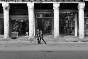 A black and white photograph of 2 young boys walking down a street with crumbling sidewalks and pillars behind them. A store advertises "Versace" and "Lacoste" in the background.