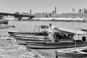 Black and white photograph of a man standing on a boat on the banks of the Tigris River. The city of Baghdad can be seen in the background.