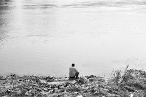A black and white photograph of a man sitting by himself on the banks of the Tigris River. The ground along the river is strewn with garbage (empty plastic bottles and debris). 