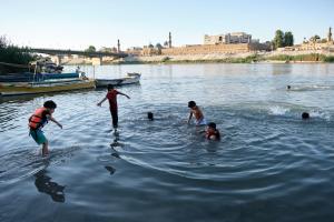 A color photograph of seven boys playing in the water of the Tigris River. The city of Baghdad can be seen in the background.