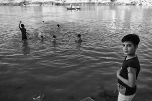 Black and white photograph of children playing in the Tigris River. A boy in the foreground stands on the banks of the river and looks directly at the camera. Five other boys play in the water and a boat with men can be seen in the background.