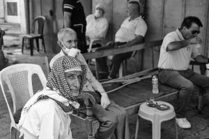 A man with a head scarf looks directly at the camera. Several other men in the background drink tea and talk, some are wearing face masks since the photo was taken during the COVID 19 pandemic 