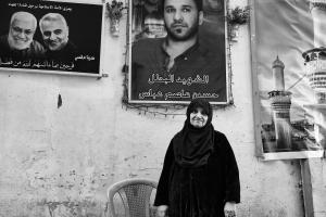 An elderly woman in traditional Iraqi clothing stands in front of a poster with a photo of her dead son 
