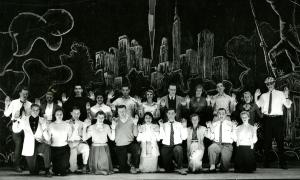 Black and white photograph of the cast of the 1956 Senior Show "Banned in Boston." The cast includes both men and women. They are assembled on a stage with a drawing of a cityscape behind them. They are arranged in two rows with the front know kneeling and they have their hands raised as if waving.