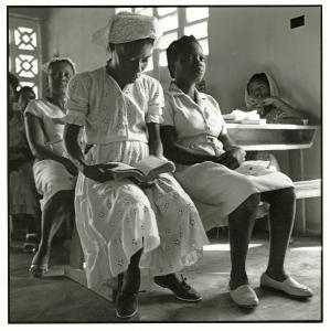 Haitian women seated, one reading book, wearing white dresses.