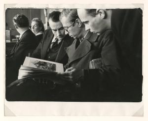 Three men standing together reading, pictures in book, wearing suits.