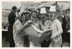Women standing in street reading among crowdn, huddled together, wearing head pieces and smiling.