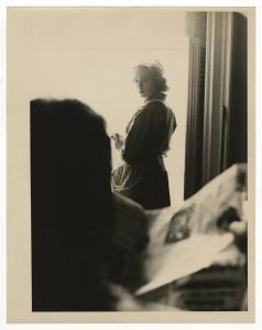 Woman standing in window, wearing apron, looking back at man reading newspaper. 
