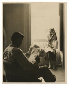 Woman seated in window leaning on broomstick, looking at man seated in chair reading newspaper.